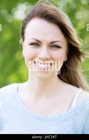 Porträt einer schönen Frau. Sie steht im Freien mit ihrem Haar im Wind und Bäume sind unscharf im Hintergrund. Stockfoto