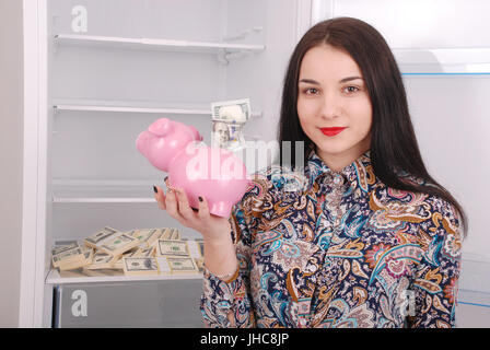 Junge schöne Frau mit Sparschwein (Spardose) auf dem Kühlschrank Hintergrund Stockfoto