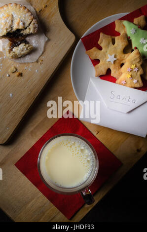 Kekse (Cookies), Milch und Mince Pie ausgelassen auf Holztisch für Santa Claus mit Umschlag mit Wunschliste.  Mince Pie ist wie du aufgebrochen Stockfoto