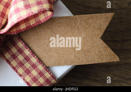 Nahaufnahme (Makro) der eine leere braune Geschenkanhänger auf einer weißen Geschenkbox mit Gingham-Band zu einer Schleife gebunden.  Eiche Holz Tischfläche unten in soft-Fokus. Stockfoto