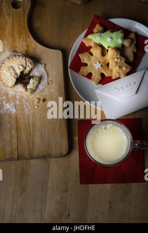 Overhead Schuss auf Holztisch Kekse (Cookies), Milch und Mince Pies, ausgelassen am Heiligabend für Santa, mit Liste in Umschlag. Stockfoto