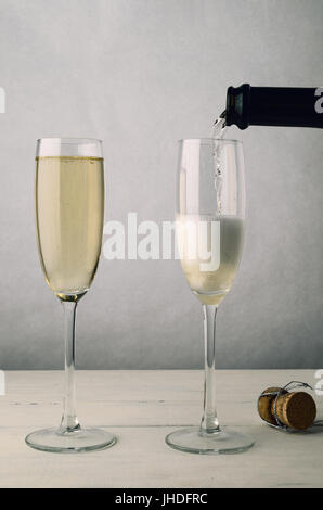 Sekt gießt aus der Flasche zu einem der zwei Sektgläser auf eine alte bemalte Holzbohle Tabelle. Das zweite Glas ist bereits voll. W Stockfoto