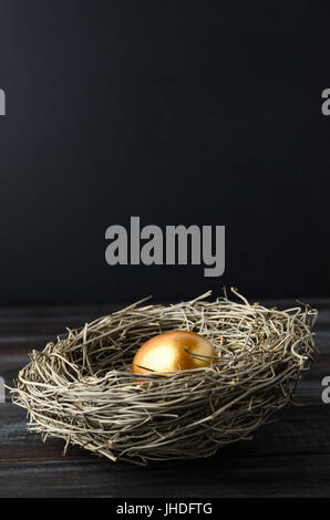 Ein einziges gold bemalten Ei in ein Vogelnest auf dunklen Holzbohle Tisch mit schwarzer Tafel Hintergrund Kopie Raum oben bieten. Stockfoto