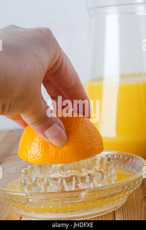 Hand greift in Rahmen von links, halben Orange auf Glas manuelle Obst Saftpresse auspressen.  Krug mit frisch gepresstem Orangensaft im Hintergrund auf hellen Holz pl Stockfoto