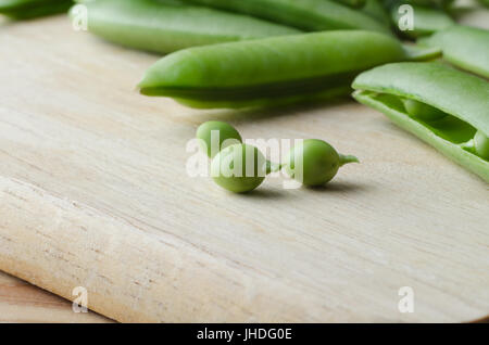 Food-Vorbereitung-Szene. Drei frisch geerntete Erbsen mit Stielen auf ein Schneidbrett aus Holz mit geöffneten und geschlossenen grünen Hülsen verstreut hinter ihnen. Stockfoto