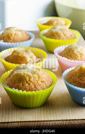 Reihen von Cupcakes, frisch aus dem Ofen, in bunten Silikon Backen Tassen auf einem Holzbrett mit Rührschüssel im Hintergrund. Stockfoto