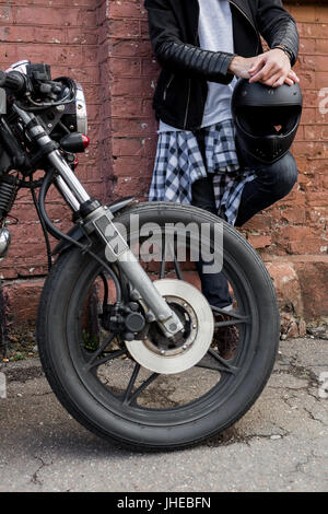 Nahaufnahme einer schönen Rider Biker Kerl Hand mit schwarzen Helm in der Nähe von Mauer und klassischen Stil Café Racer Motorrad. Fahrrad benutzerdefinierte gemacht in Vintage gara Stockfoto