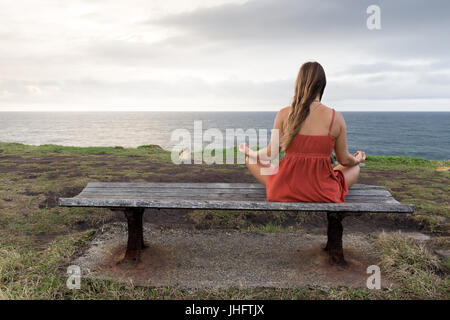 Eine Frau in einem orange Kleid meditiert auf einer Holzbank am Meer. Stockfoto