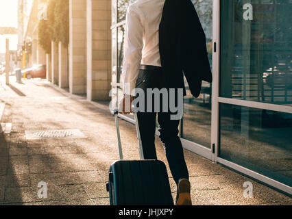 Rückansicht der junge Geschäftsmann zu Fuß außerhalb ÖPNV Gebäude mit Koffer. Business Traveller Koffer an Stadt Straße ziehen. Stockfoto