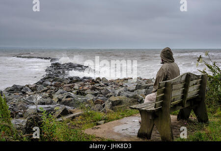 Einzelperson im Urlaub sitzt auf einer Bank mit Blick auf ein raues Meer und Sturmwolken. Stockfoto