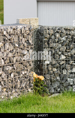 graue Gabione mit Kies, Steinen und Steinplatten vor grünen Rasen Stockfoto