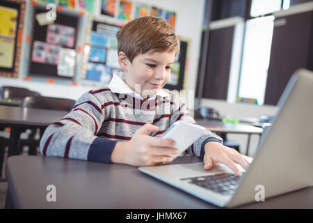 Lächelnde Schuljunge mit Laptop und Handy am Schreibtisch im Klassenzimmer Stockfoto