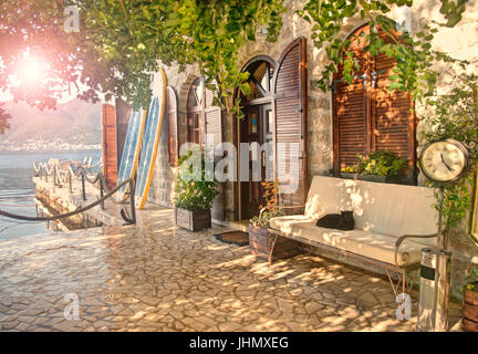 warmen Crossen Bild der Sonne durch Blätter von Jahrhunderte alten Johannisbrotbaum neben alten Hauseingang mit Surfbretter, Bank, Katze liegend auf Sie Stockfoto