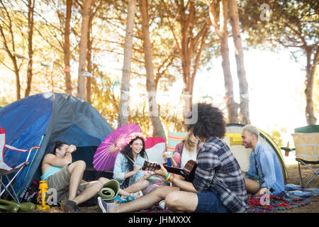 Mann, die Gitarre zu spielen, während Sie sitzen mit Freunden auf Feld auf Campingplatz Stockfoto