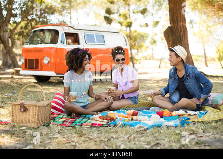 Lächelnd Freunde etwas zu Essen beim Sitzen auf der Picknickdecke im Feld gegen Wohnmobil Stockfoto