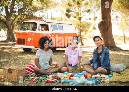 Porträt von lächelnden Freunde etwas zu Essen beim Sitzen auf der Picknickdecke im Feld gegen Wohnmobil Stockfoto