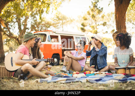 Mann Fotografieren Freunde beim Sitzen auf auffangene gegen Wohnmobil bei Picknick Stockfoto