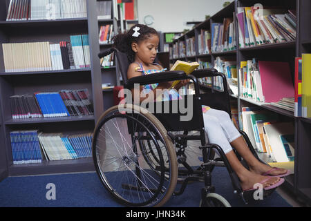 Mädchen lesen Buch beim Sitzen im Rollstuhl durch Regale in Bibliothek Stockfoto