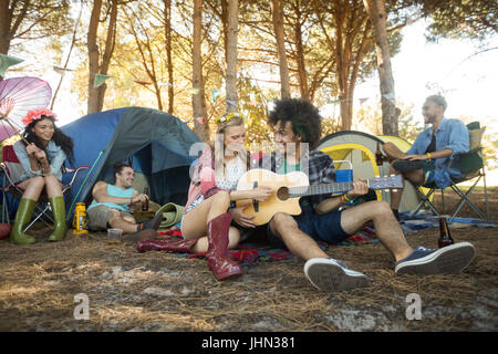 Junge Frau mit männlichen Freund Gitarre sitzend auf Feld auf Campingplatz Stockfoto