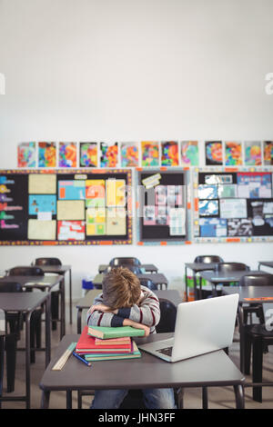 Müde Schüler schlafen auf Stapel von Buch von Laptop beim Sitzen am Schreibtisch im Klassenzimmer Stockfoto