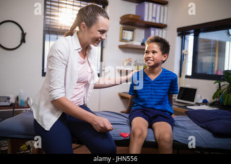 Lächelnde weiblichen Therapeuten Blick auf jungen beim schlagen am Knie mit Reflexhammer auf Krankenstation Stockfoto