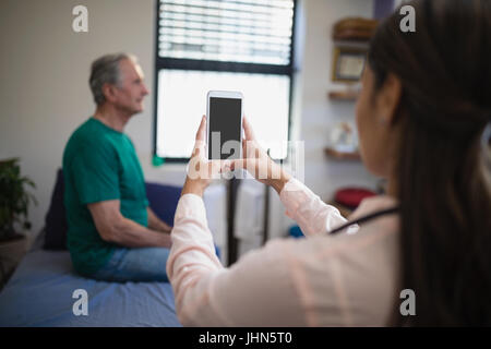 Rückansicht des weiblichen Therapeuten fotografieren senior männlichen Patienten von Handy gegen Fenster auf Krankenstation Stockfoto