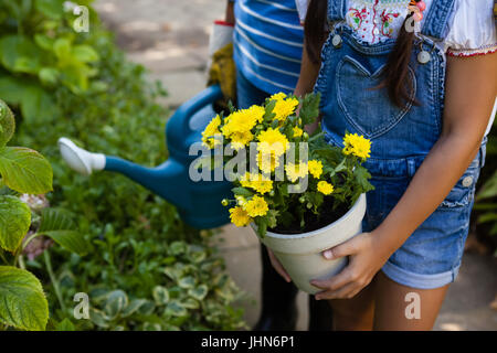 Mittelteil der älteren Frau und Enkelin mit Gießkanne und gelben Blumentopf im Hinterhof Stockfoto