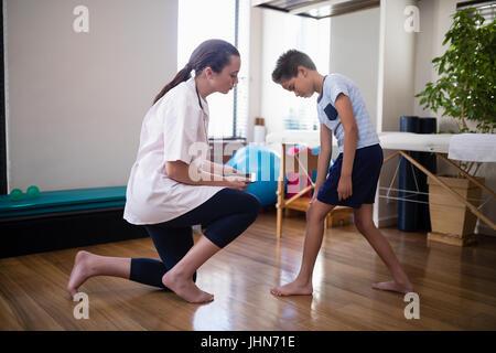 Jungen zeigen Knie zum weiblichen Therapeuten kniend auf Hartholz-Fußboden im Krankenhaus Stockfoto