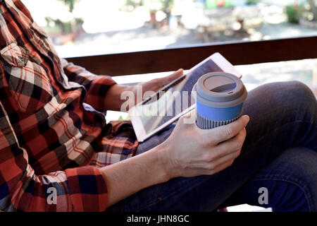 Closeup von einem kaukasischen Jüngling trägt Jeans und ein kariertes Hemd mit seinem Tablet auf der Veranda eines Hauses oder einer ranch Stockfoto