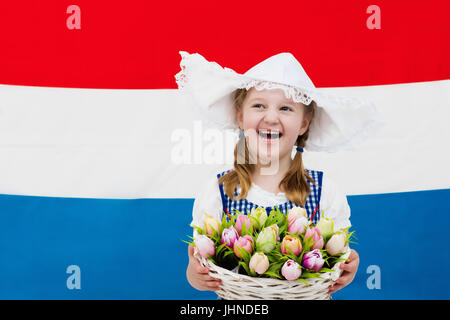 Niederländische Mädchen tragen traditionelle Tracht, Kleid und Hut hält Korb mit Tulpen auf der Flagge der Niederlande. Kind mit Souvenirs aus H Stockfoto