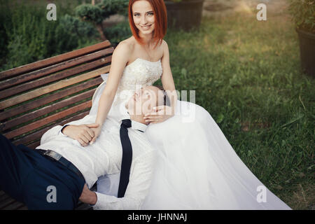 Bräutigam liegt auf das schöne Mädchen in einem Brautkleid im öffentlichen Park auf der Bank. Braut ist Rothaarige legte eine Hand auf die Msn und lächelnd Stockfoto