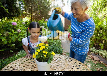 Mädchen auf der Suche während lächelnd senior Frau Bewässerung gelbe Blumen auf Tisch im Garten Stockfoto