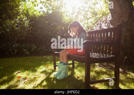 Lächelndes Mädchen mit Smartphone beim Sitzen auf der Holzbank im Hinterhof an sonnigen Tag Stockfoto