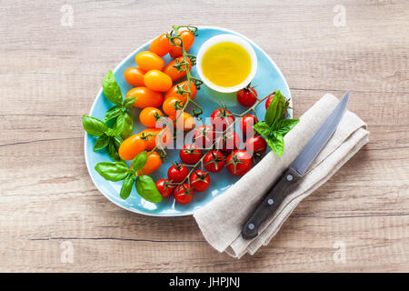 Vielzahl von Cherry-Tomaten und Basilikum Blätter auf der blauen Platte auf einem Holztisch. Zutaten für gesunde Sommersalat Stockfoto