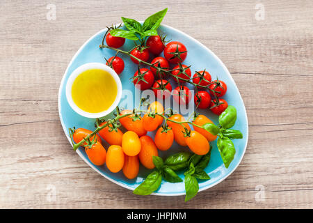 Vielzahl von Cherry-Tomaten und Basilikum Blätter auf der blauen Platte auf einem Holztisch. Zutaten für gesunde Sommersalat Stockfoto