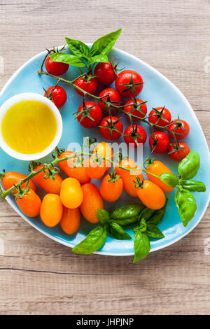 Vielzahl von Cherry-Tomaten und Basilikum Blätter auf der blauen Platte auf einem Holztisch. Zutaten für gesunde Sommersalat Stockfoto
