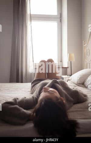 Junge Frau am Bett gegen Fenster zu Hause entspannen Stockfoto