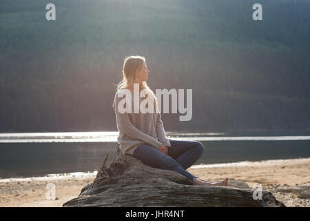 Junge Frau sitzend auf Baumstamm und Wegschauen am Seeufer Stockfoto