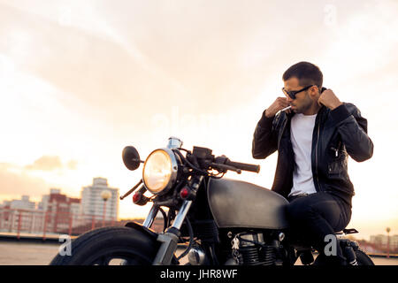 Hübscher Reiter Mann mit Bart und Schnurrbart in schwarze Mode Sonnenbrillen Rauchen Strafpredigt und richtige Biker Jacke sitzen auf klassischen Stil Café Racer motorb Stockfoto