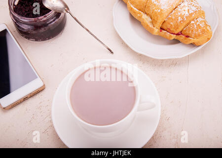 Croissant mit Himbeermarmelade, Kakao in einem Becher in der Hand Handy. Business-Frühstück-Konzept. Stockfoto