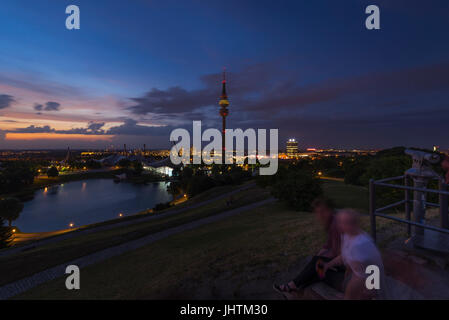 Leute sitzen auf einem Hügel im Olympischen Park beobachten Sonnenuntergang über olympische lak, Olympiaturm in München Stockfoto