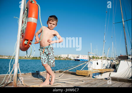 lustige kleine gesunde junge in kurzen Hosen steht auf dem Pier mit Meer und blauer Himmelshintergrund Stockfoto