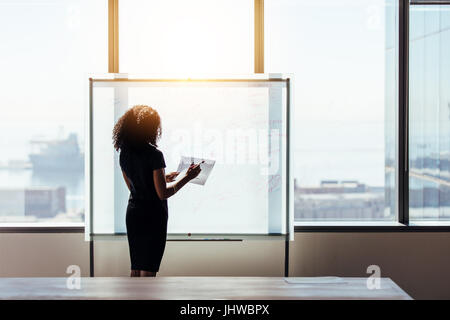 Heckansicht des eine Geschäftsfrau auf einem Whiteboard im Büro schreiben. Unternehmen Investor halten Sie Papier und Stift stand vor eine Präsentationstafel. Stockfoto