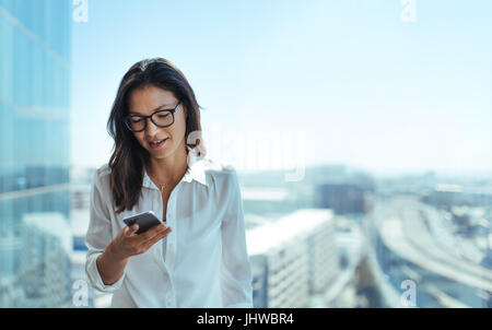 Geschäftsfrau Kurzmitteilungen über Handy. Junge Frau in ihrem Büro in einem Hochhaus mit Blick auf das Stadtbild. Stockfoto