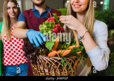 Der Verkäufer und zwei Mädchen mit einem Korb voller Gemüse. Freundinnen kaufen Gemüse auf dem Markt. Stockfoto