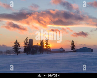 Ein farbenfrohen Sonnenuntergang hinter Wirtschaftsgebäude beleuchtet Wellen der Schnee weht über ein Feld im Vordergrund. Stockfoto