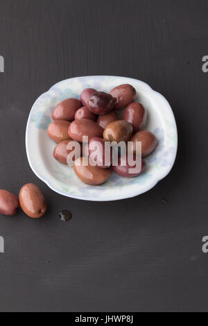 Violette Oliven in kleine weiße Schüssel mit zwei auf schwarzer Kreide Board Textur Hintergrund - vertikale Tageslicht schoss mit Platz für text Stockfoto