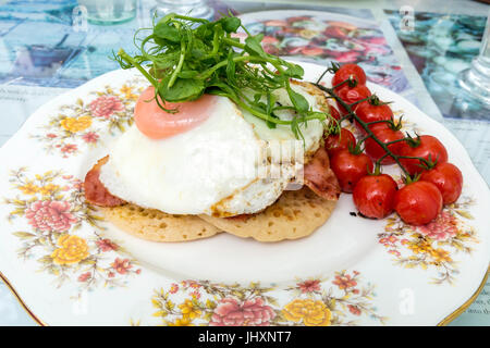 Ein leckeres Mittagessen mit zwei Spiegeleier und Speck auf Pikelets mit Cherry-Tomaten und Wasser Kresse garnieren in einem Café in North Yorkshire Stockfoto