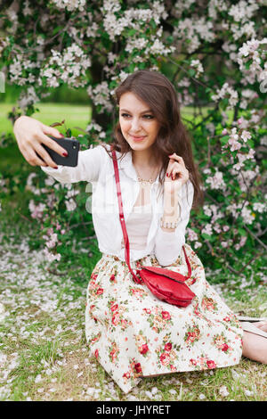 Ziemlich lächelnde junge Frau mit dem Handy telefonieren, Fotografieren im Freien. Frau in einem weißen Pullover und blaue Jeans macht Selfie sitzen auf dem Rasen und f Stockfoto