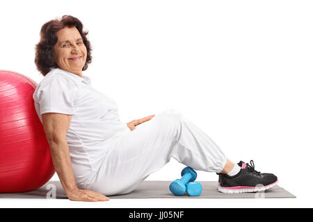 Reife Frau mit einem Pilates-Ball und Kurzhanteln, sitzend auf einer Ausübung Matte isoliert auf weißem Hintergrund Stockfoto